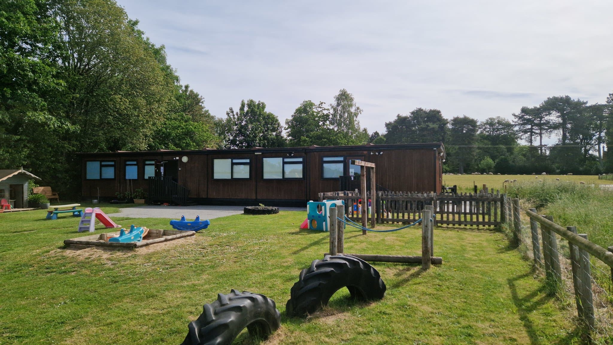 Bojangles Day Nursery outdoor play area with a sandpit, see-saw, climbing frame and tractor tyres on the grass in front of the nursery building